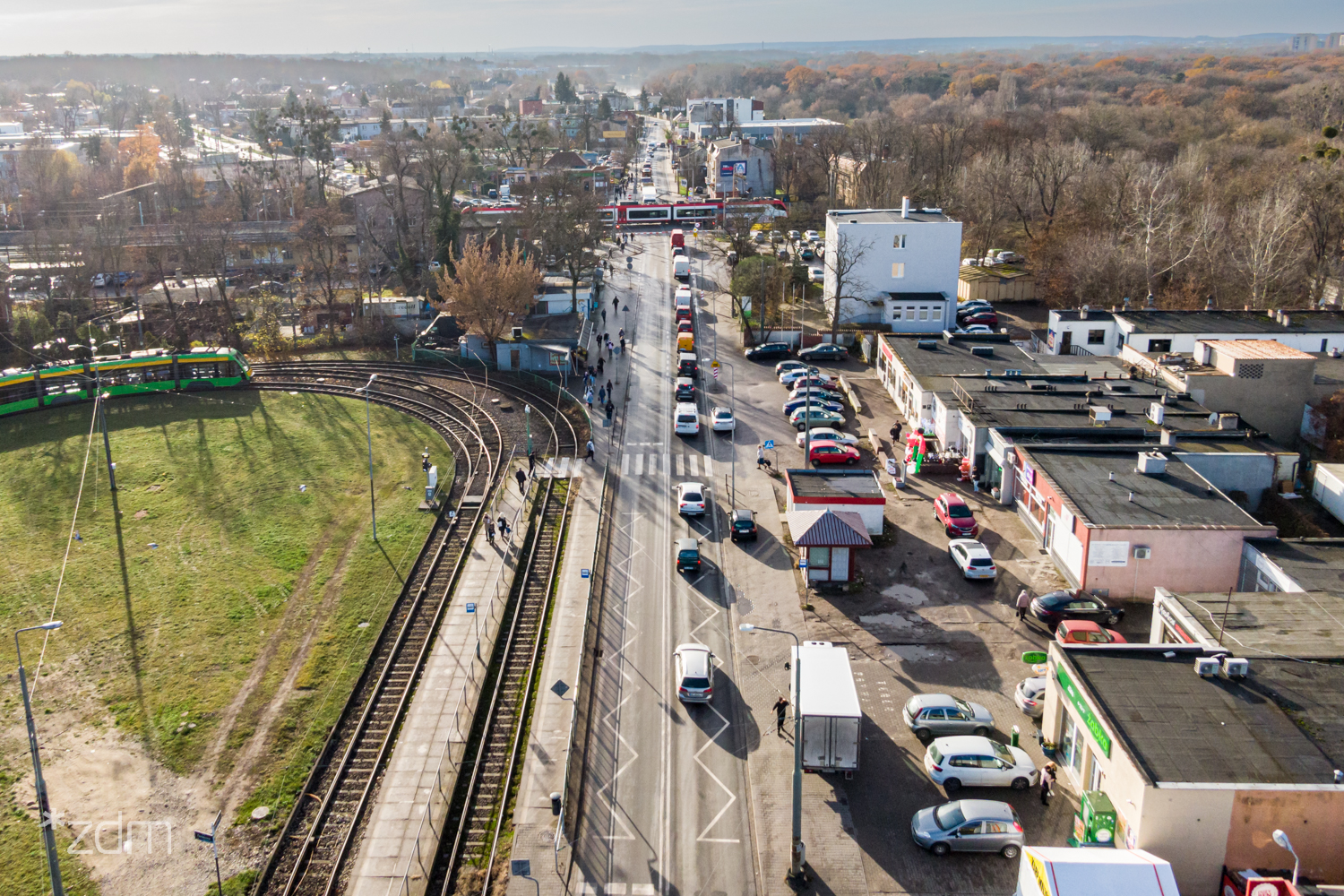 Widok z lotu ptaka na zamknięty przejazd kolejowy przy ul. Starołęckiej, przez który przejeżdża pociąg. Samochody czekają na otwracie szlabanów. Po prawej stronie jest pętla tramwajowa - grafika artykułu