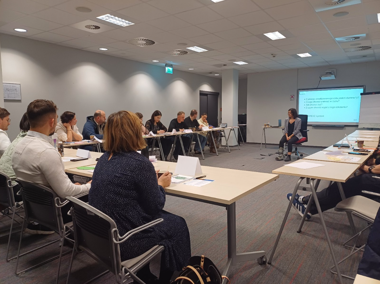 Grupa ludzi siedzi wokół stołu ustawionego w kształt litery U w sali konferencyjnej. Jeden z uczestników prowadzi prezentację lub dyskusję, stojąc lub siedząc na przedzie pomieszczenia. - grafika artykułu