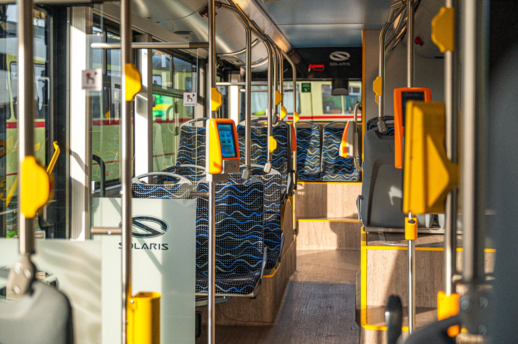 Wnętrze autobusu marki Solaris. Autobus charakteryzuje się nowoczesnym designem, w tym wzorzystymi siedzeniami w niebiesko-czarne fale, żółtymi poręczami oraz pomarańczowymi kasownikami biletów. Logo Solaris jest widoczne na przegrodzie w pobliżu przedniej części autobusu. Wnętrze jest dobrze oświetlone dzięki naturalnemu światłu wpadającemu przez okna. - grafika artykułu