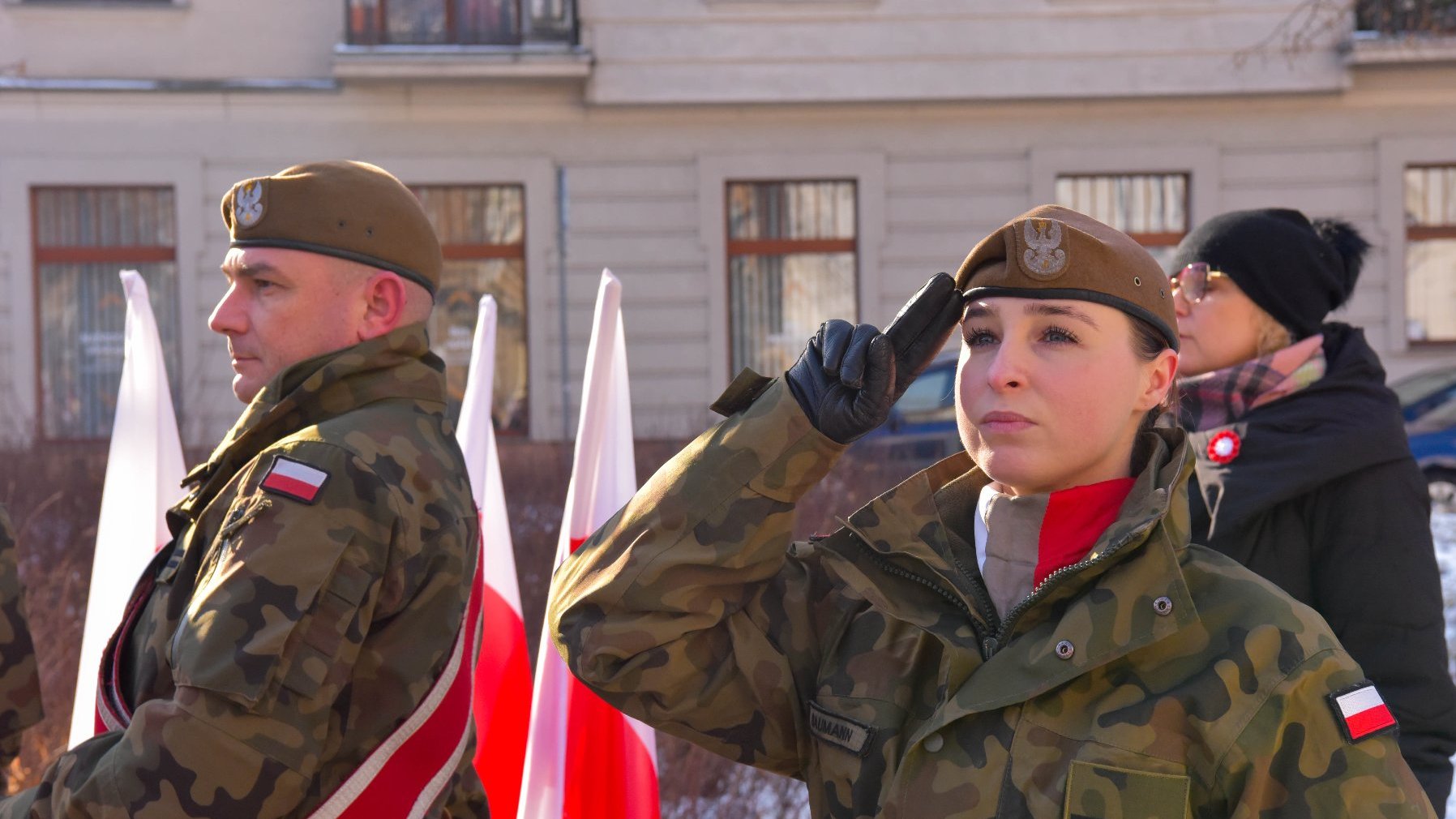 Dwóch mężczyzn w mundurach wojskowych stoi na zewnątrz. Obaj mają na sobie kurtki w kamuflażu i berety z odznakami. Mężczyzna po prawej stronie salutuje prawą ręką, mając na sobie czarną rękawiczkę. Na lewym ramieniu obu mężczyzn znajduje się naszywka z polską flagą. W tle widoczne są polskie flagi oraz budynek z wieloma oknami.