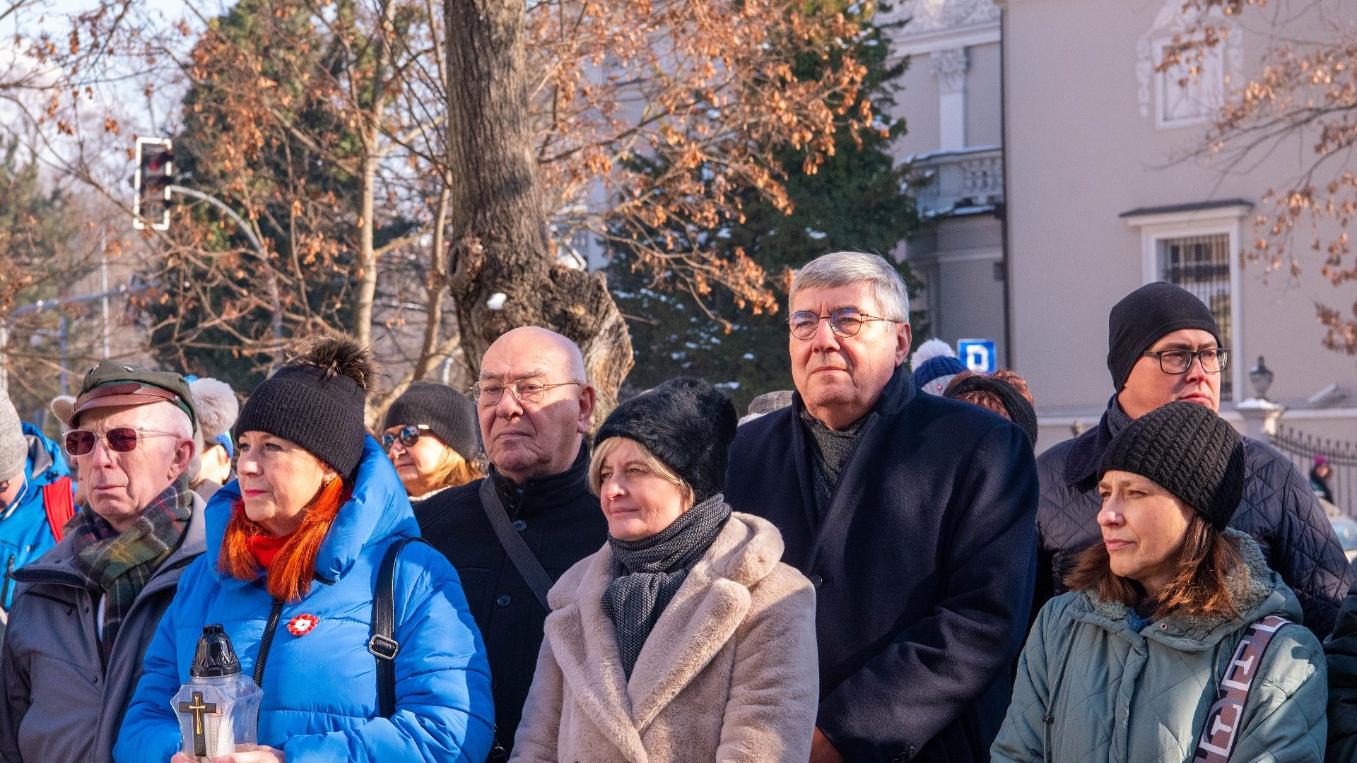 Grupa ludzi stojących na zewnątrz podczas wydarzenia. Ubrani są w zimowe płaszcze, szaliki i czapki, co wskazuje na chłodną pogodę. Na pierwszym planie osoba trzymająca latarnię z krzyżem. W tle drzewa z rzadkimi liśćmi i budynek z detalami architektonicznymi.