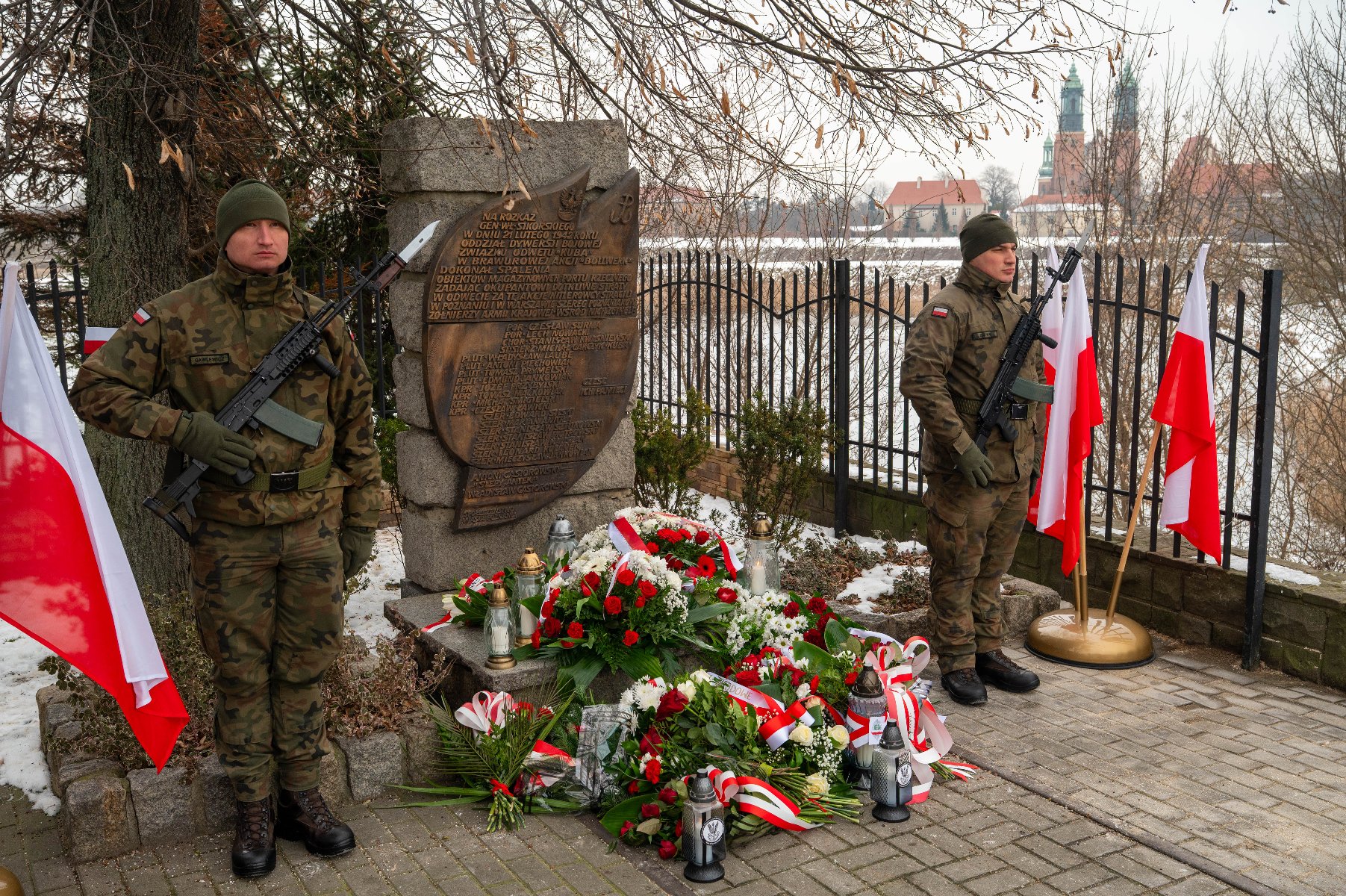 Zdjęcie przedstawia dwóch żołnierzy stojących na straży obok pomnika pamiątkowego. Żołnierze są ubrani w mundury kamuflażowe i trzymają karabiny. Pomnik wykonany jest z kamienia i ma wyryty napis na tabliczce. Wokół pomnika znajduje się wiele kwiatowych wieńców i bukietów, z których wiele jest ozdobionych czerwono-białymi wstążkami, co jest nawiązaniem do kolorów flagi Polski. Obok pomnika ustawiono również kilka flag Polski. - grafika artykułu