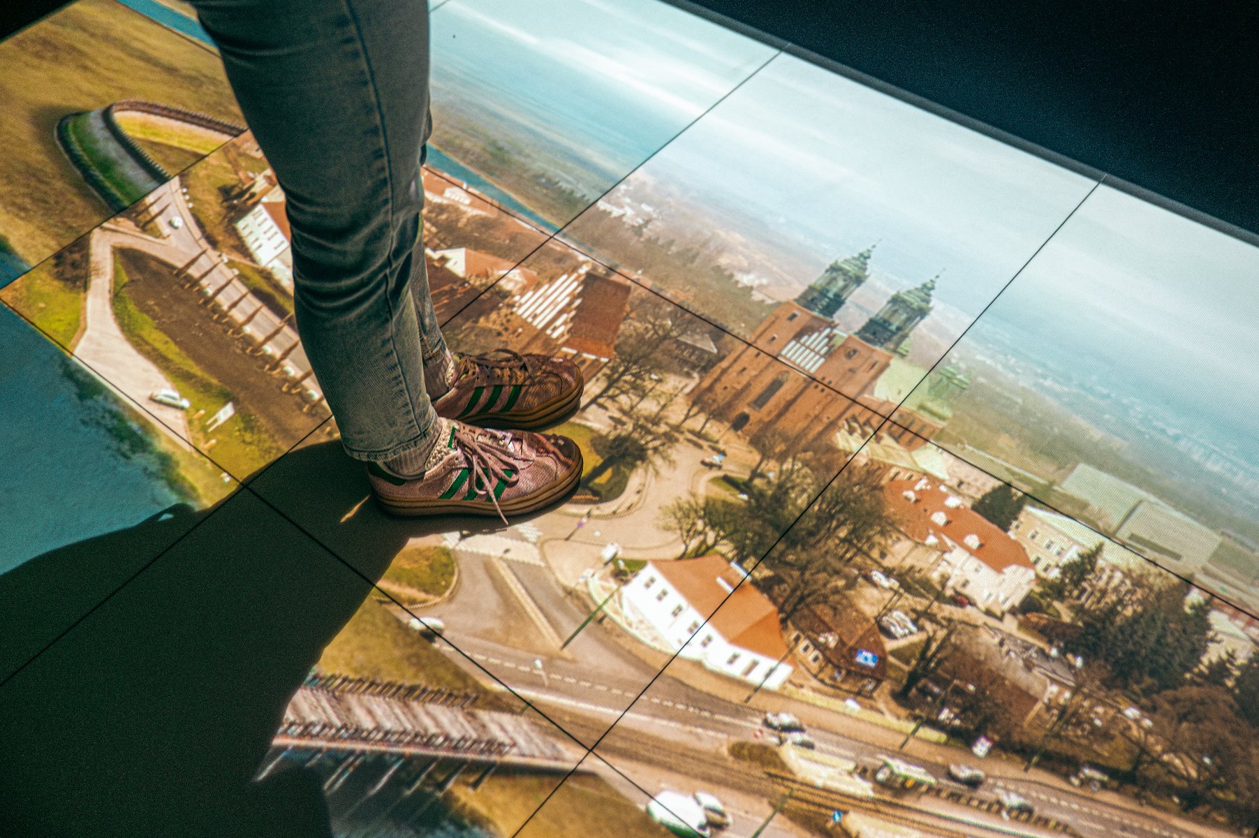 Zdjęcie przedstawia fotografię Ostrowa Tumskiego, m.in. katedry poznańskiej na podłodze oraz nogi osoby, kóra na nim stoi. - grafika artykułu