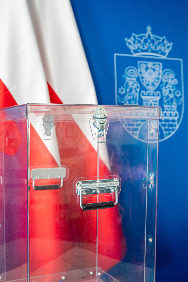 Zdjęcie przeźroczystej urny wyborczej na tle biało-czerwonych flag i godła Miasta Poznania na niebieskim tle. - grafika artykułu