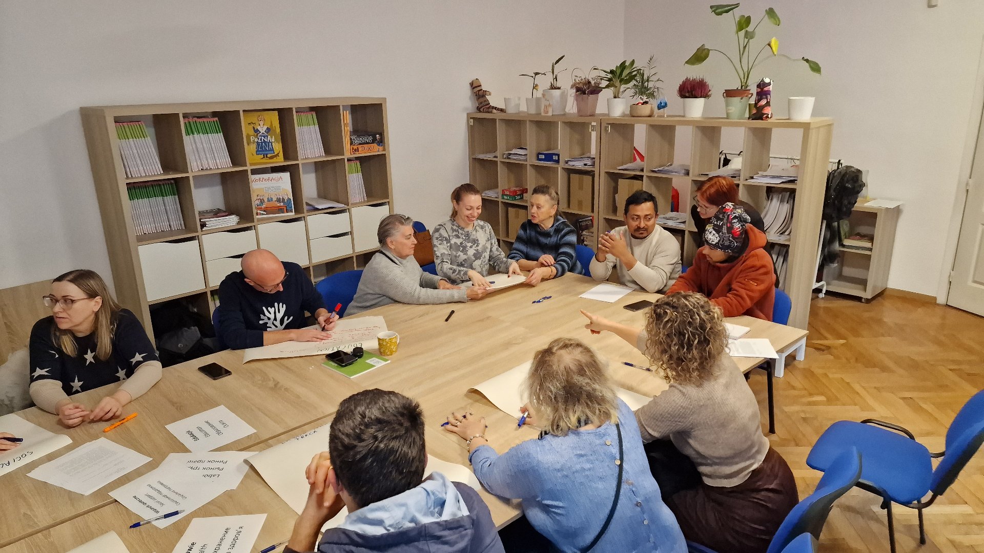 Sala warsztatowa, wokół dużego stołu siedzi grupa wielu ludzi. Na stole leżą dokumenty i papiery. Grupa rozmawia i pracuje nad dokumentami. W tle regały z ksiązkami. - grafika artykułu