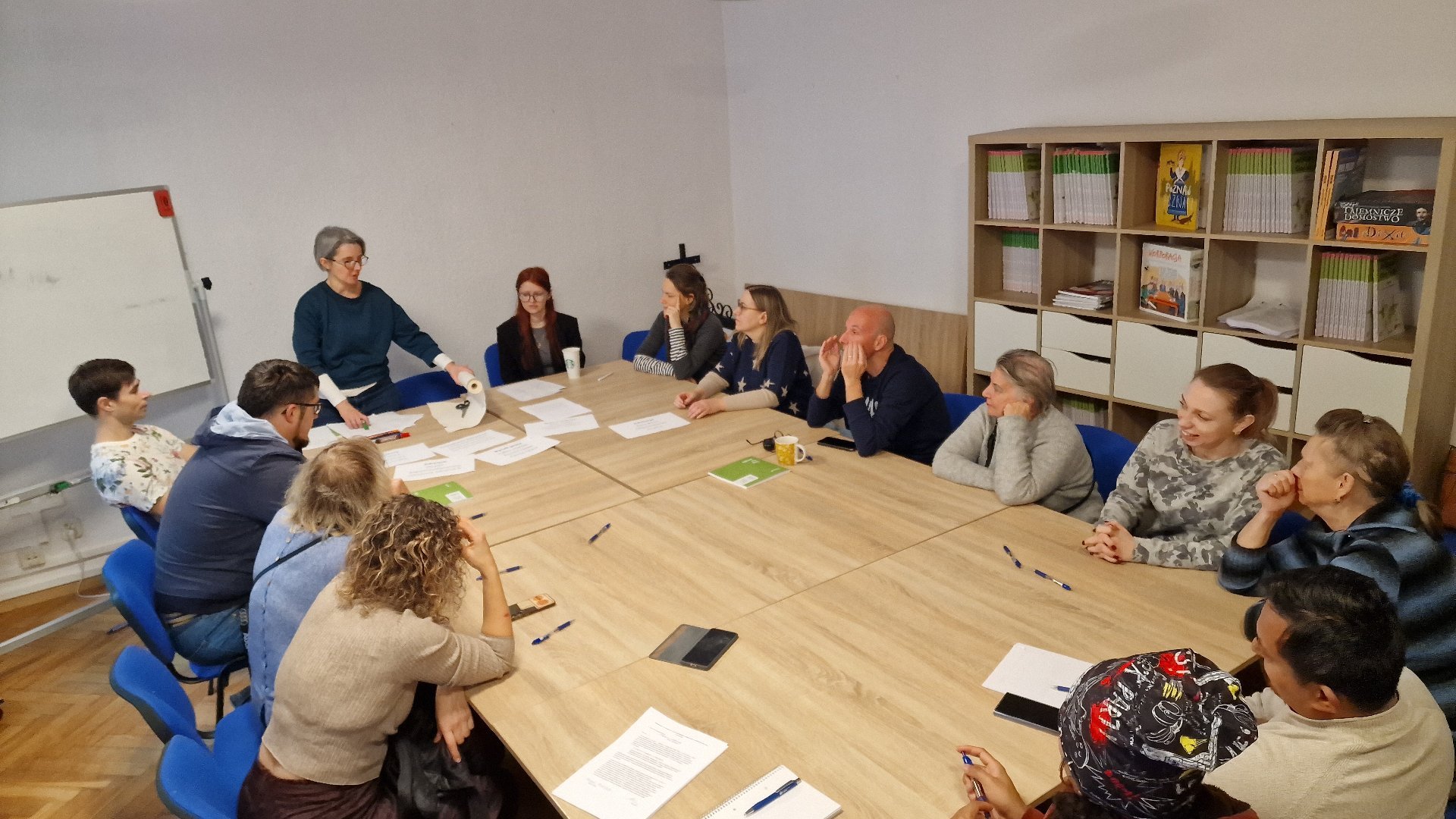 Sala warsztatowa, wokół dużego stołu siedzi grupa wielu ludzi. Na stole leżą dokumenty i papiery. Grupa rozmawia i pracuje nad dokumentami. W tle regały z ksiązkami.