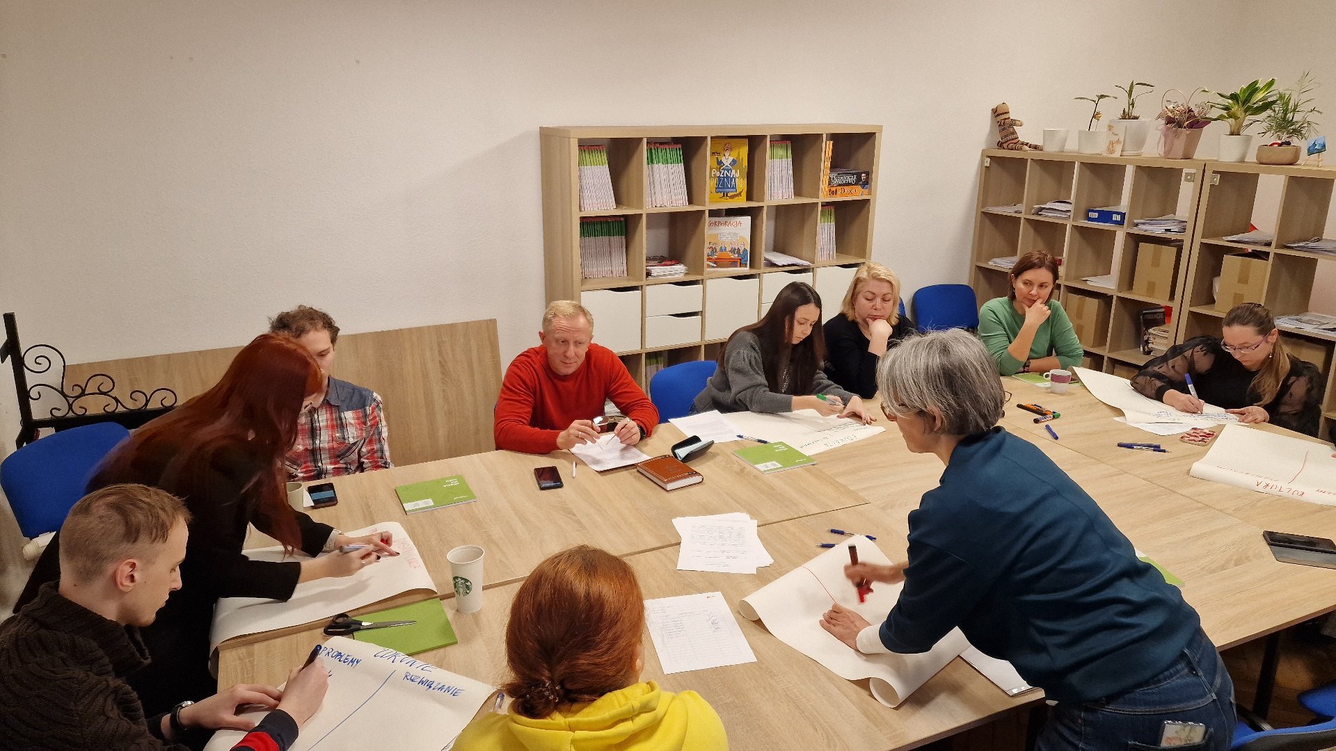 Sala warsztatowa, wokół dużego stołu siedzi grupa wielu ludzi. Na stole leżą dokumenty i papiery. Grupa rozmawia i pracuje nad dokumentami. W tle regały z ksiązkami.