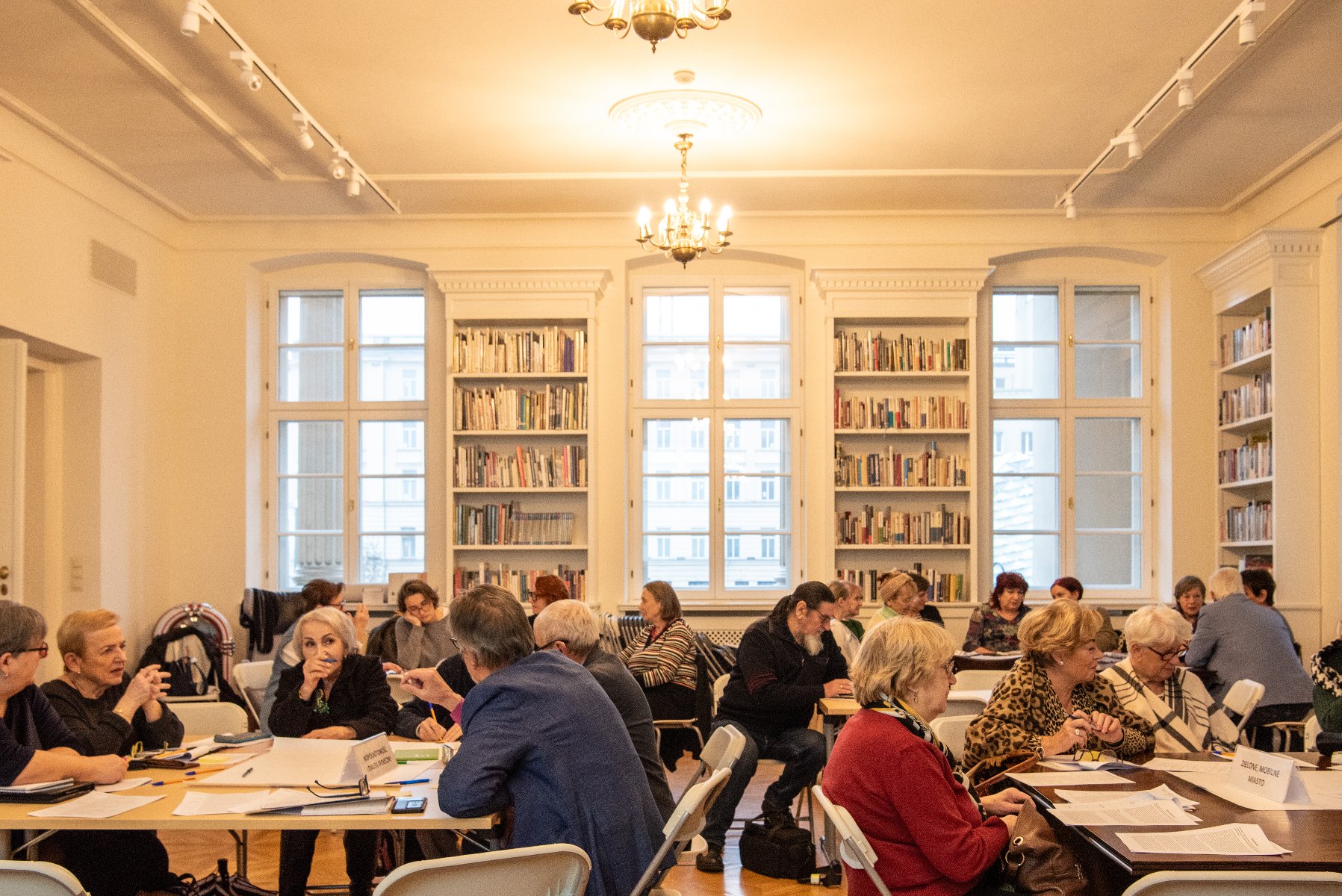 duża jasna sala biblioteki z dużą liczbą stolików, przy których siedzą seniorki i seniorzy. Trwają warsztaty i praca w grupach. - grafika artykułu