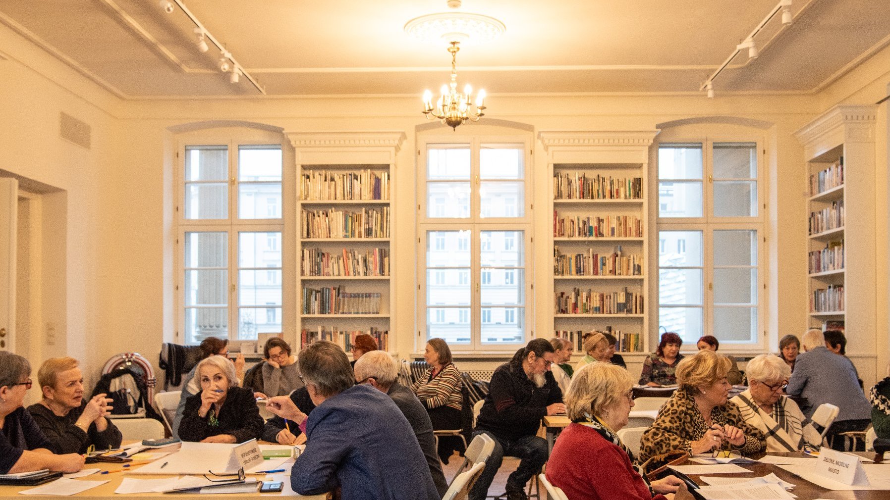 duża jasna sala biblioteki z dużą liczbą stolików, przy których siedzą seniorki i seniorzy. Trwają warsztaty i praca w grupach.