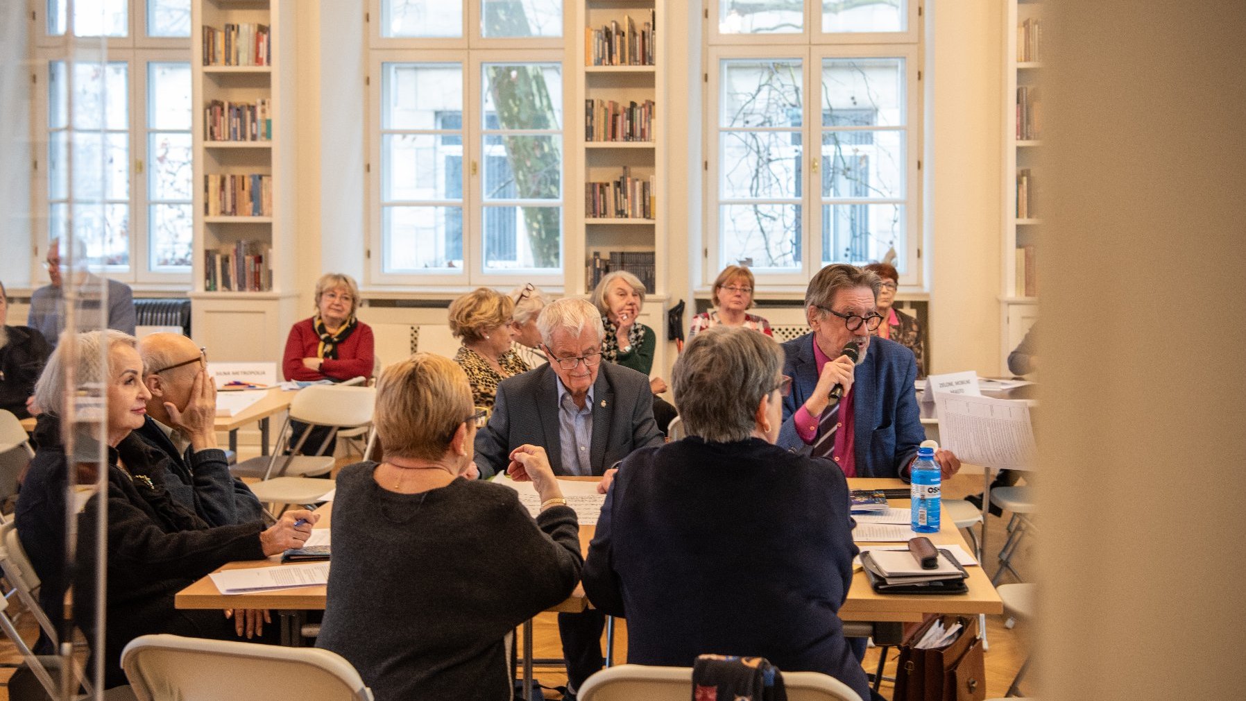 duża jasna sala biblioteki z dużą liczbą stolików, przy których siedzą seniorki i seniorzy. Trwają warsztaty i praca w grupach.