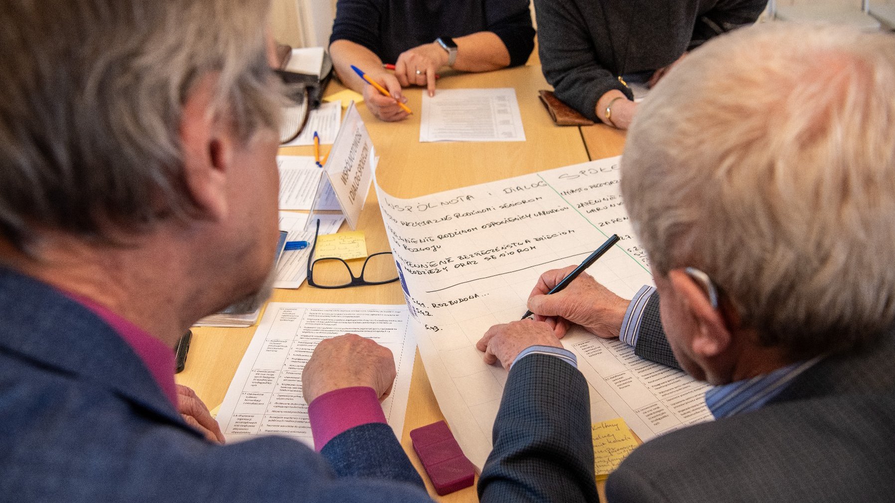 widok z lotu ptaka na pracującą grupę seniorów, na stole leżą kartki, okulary i długopisy. Jeden z seniorów zapisuje coś na dużym arkuszu papieru.