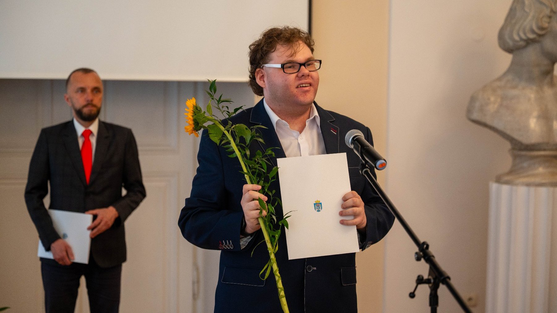 Dwie osoby stoją w formalnym otoczeniu, prawdopodobnie podczas ceremonii wręczenia nagród. Osoba na pierwszym planie trzyma słonecznik oraz certyfikat z herbem, stojąc przy mikrofonie.