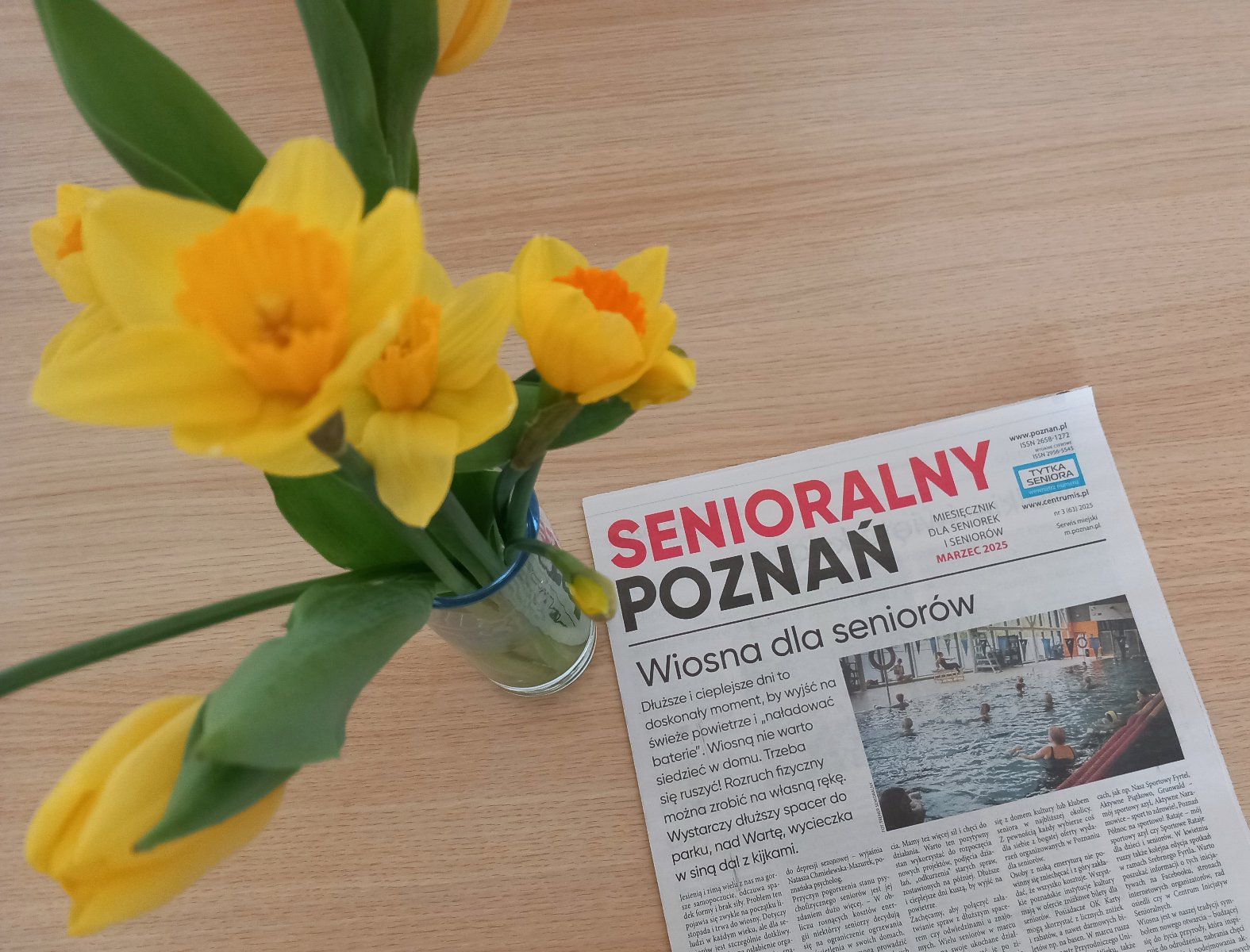 Na brązowym stoliku widać egzemplarz gazety Senioralny Poznań, obok stoi przezroczysty wazon z żonkilami - grafika artykułu