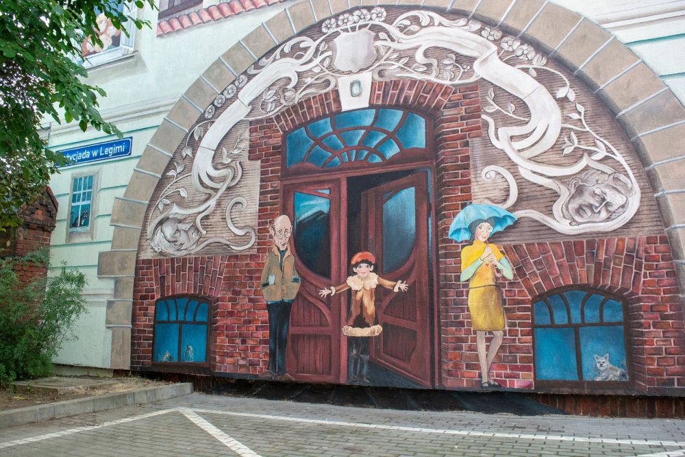 Mural - namalowane wejście do kamienicy, przy którym stoją trzy postacie z serii, starszy, siwy pan w okularach, dziewczynka w brązowym płaszczyku i czerwonym berecie oraz młoda dziewczyna w żółtej sukience. W piwnicznych okienkach widać zwierzęta, psa oraz kota. - grafika artykułu