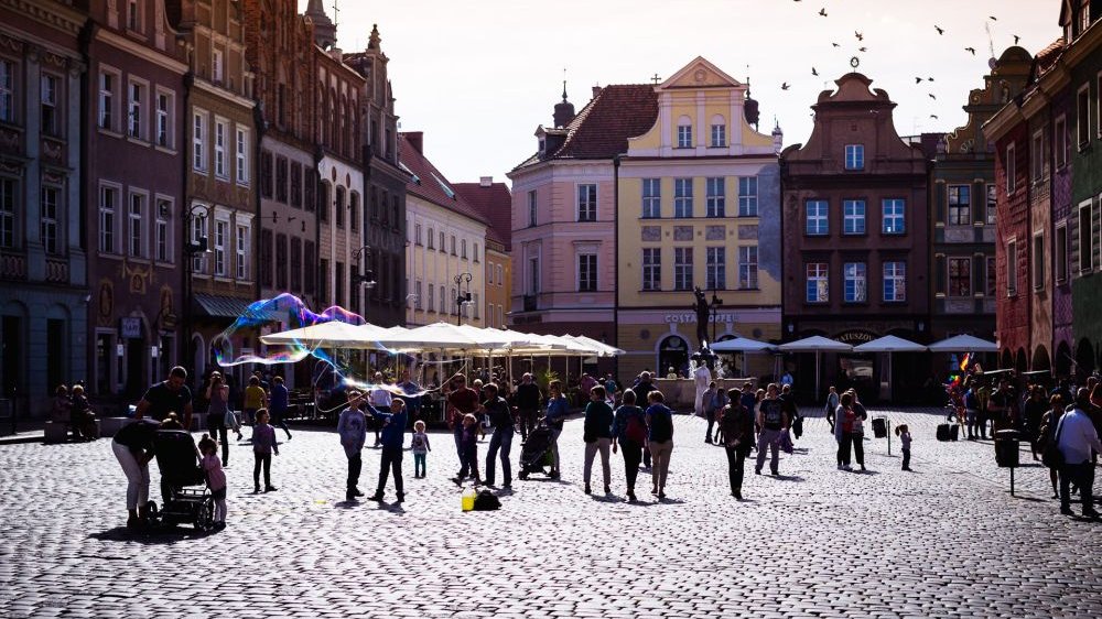 Ludzie spacerujący po Starym Rynku w Poznaniu. Kolorowe fasady kamienic. Ktoś puszcza duże bańki mydlane.