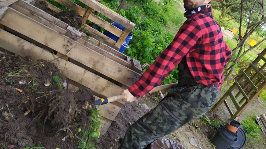 Młody mężczyzna ma czarne włosy i bródkę, jest ubrany w kraciastą koszulę i słomkowy kapelusz oraz pochylony z łopatą nad drewnianym kompostownikiem.