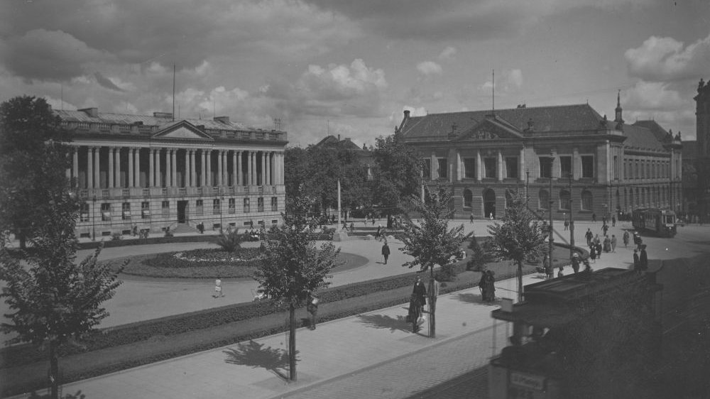 Czarno-białe zdjęcie. Fotograf, stojąc na podwyższeniu, pokazał Plac Wolności, którym przechadzają się spacerowicze. Widać budynek Biblioteki Raczyńskich i stojący po drugiej stronie ulicy gmach Muzeum Narodowego.