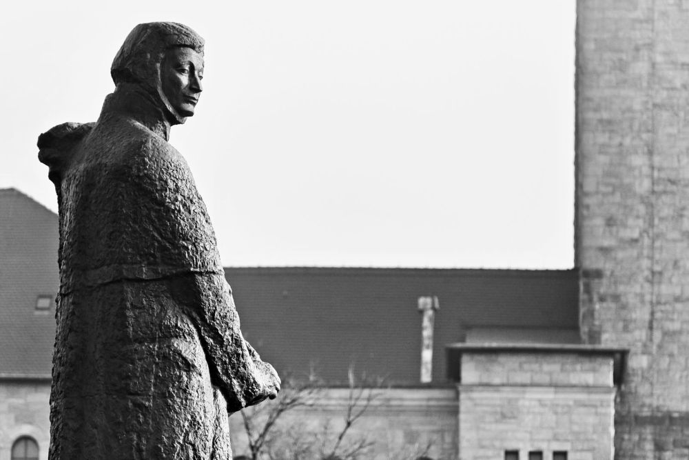 Pomnik uchwycony z profilu, Adam Mickiewicz zdaje się na nas patrzeć kątem oka. Za pomnikiem fragment murów Centrum Kultury Zamek. - grafika artykułu