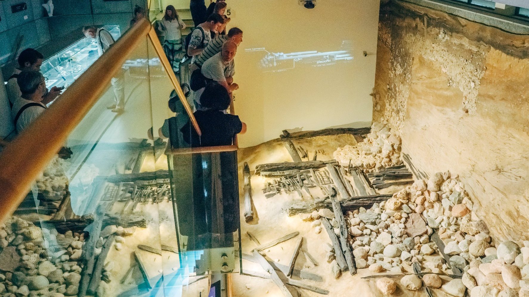 Na zdjęciu ludzie oprowadzani po ekspozycji Genius loci. Stoją na szklanym podeście, pod spodem ruiny, mury.