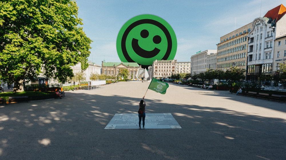 Osoba stojąca na dużym placu, macha zieloną flagą. W oddali budynki, zza których wystaje emotikon uśmiechniętej buzi.