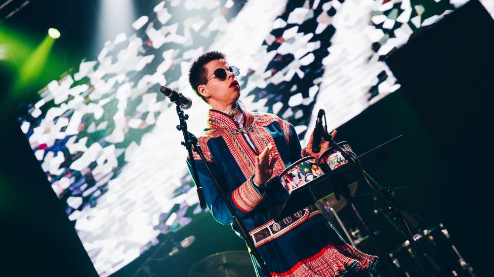 A man in a Norwegian folk costume and sunglasses plays drums on stage.