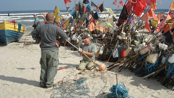 Nadmorska plaża. Dwóch mężczyzn pracuje przy łodzi rybackiej. Jeden z nich stoi tyłem, drugi klęczy. Za nimi kolorowe flagi.