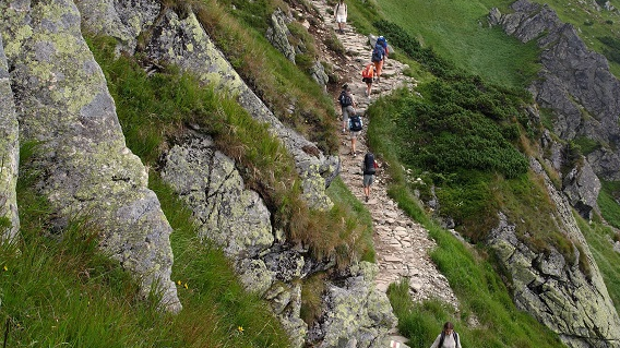 Górski krajobraz. Po zboczu góry, kamienną ścieżką idzie kilku turystów.