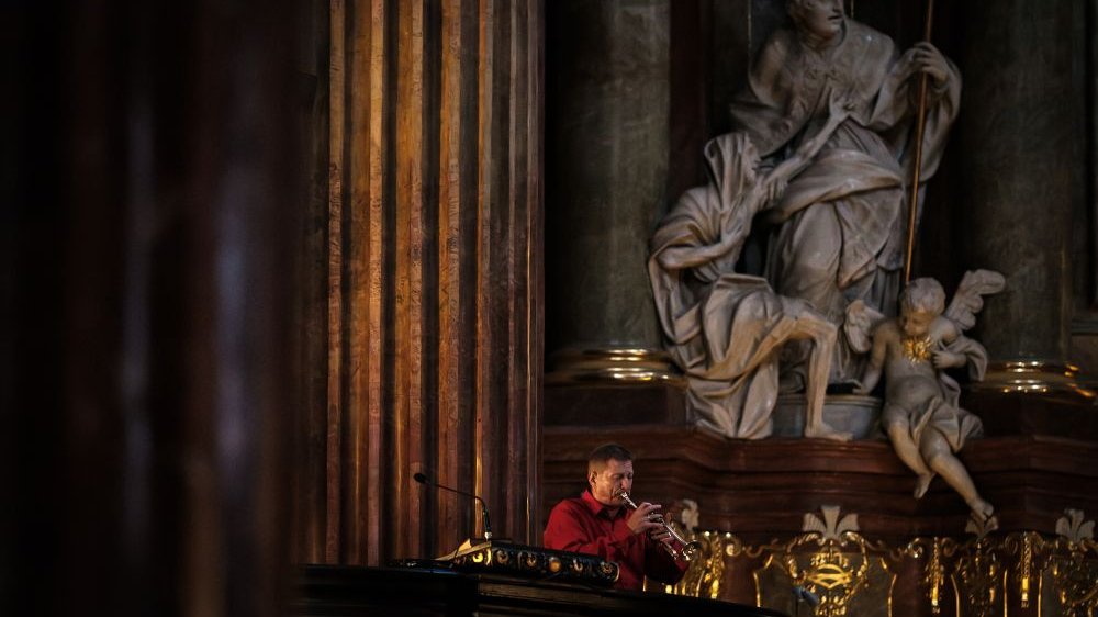 Muzyk w czerwonej koszuli gra na trąbce, stojąc z boku ołtarza. Nad nim górują rzeźby w farze, przedstawiają świętych i cherubiny.