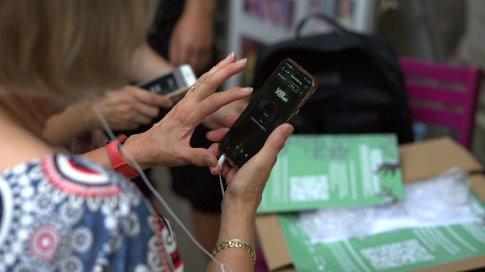 Stojąca tyłem kobieta sprawdza apliakcję na ekranie smartfona. Na stole leżą zielone plakaty.