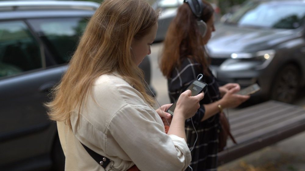 Dwie młode dziewczyny stoją na ulicy. Słuchają nagrania na słuchawkach podłączonych do swoich smartfonów.