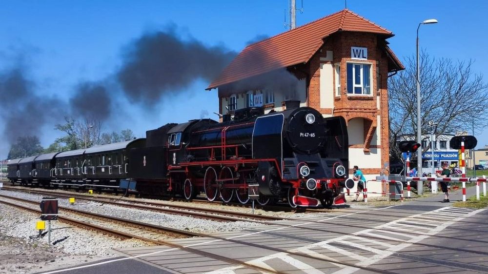 Skład ma czarną staroświecką lokomotywę i ok. pięć wagonów. Stoi na stacji w pogodny dzień, z lokomotywy bucha para.