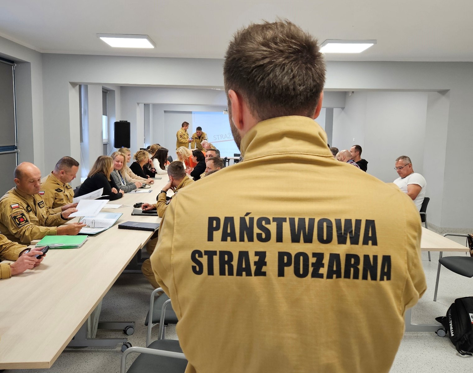 Na zdjęciu widać ludzi podczas szkolenia w sali. Na pierwszym planie jest osoba w żółtej koszulce z napisem Państwowa Straż Pożarna. Osoba ta stoi tyłem do obiektywu. W tle zdjęcia widać inne osoby, które siedzą przy stołach. - grafika artykułu