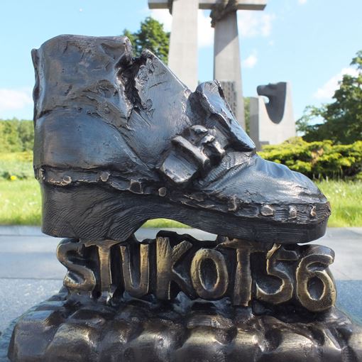 Mosiężny but z napisem "Stukot 56" pod podeszwą. W tle pomnik Trzech krzyży i zielona roślinność - grafika artykułu