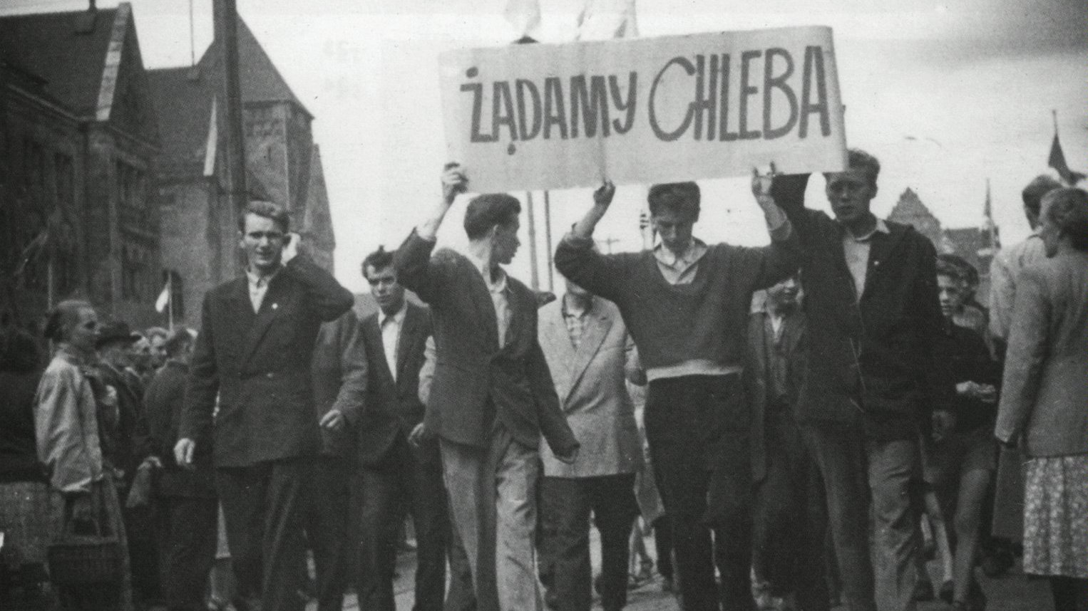 Pochód robotników. Trzech mężczyzn na przodzie trzyma transparent z napisem "Żądamy chleba". Po prawej stronie idzie Janusz Kulas.