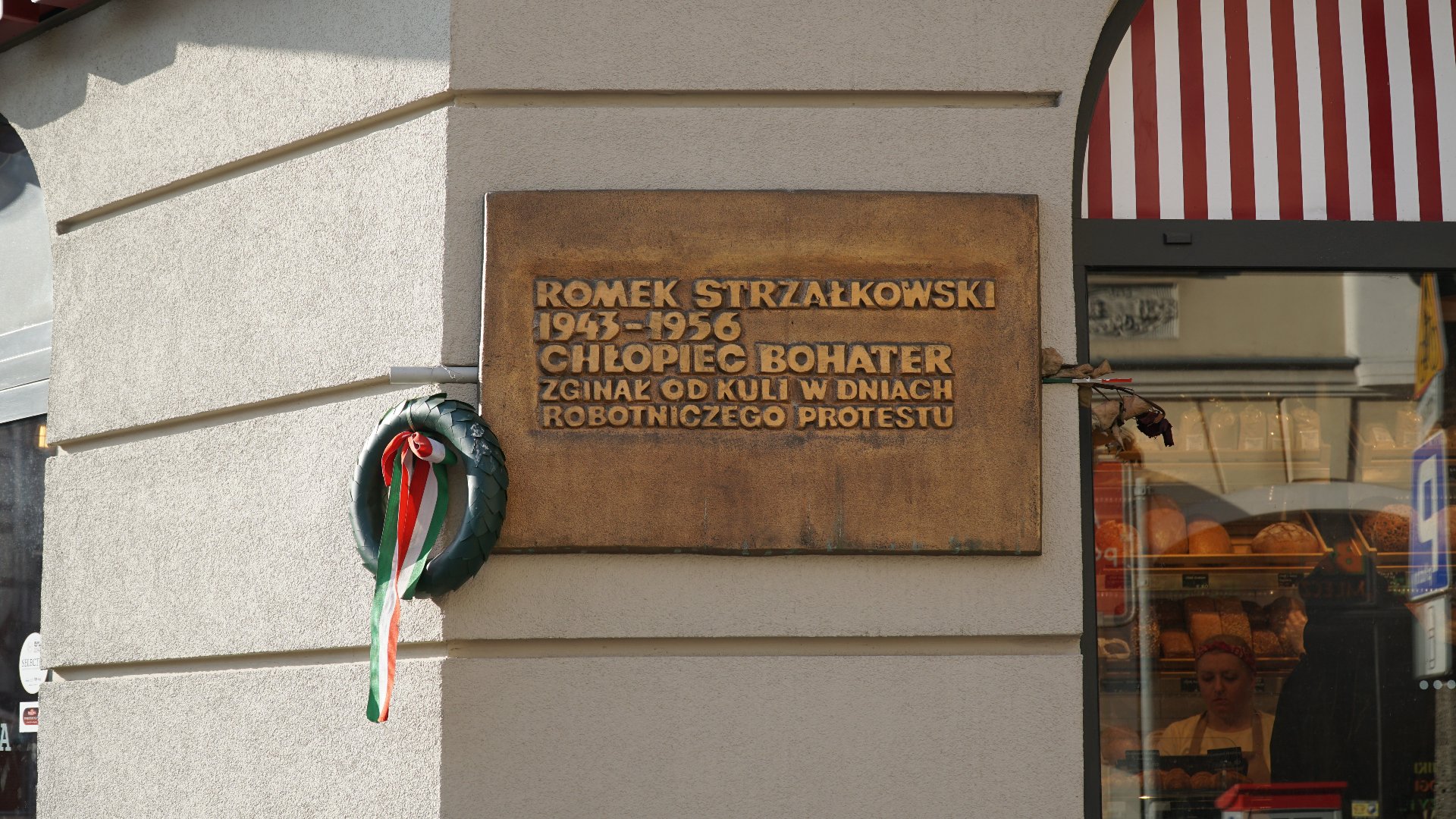 Miedziana tablica powieszona na rogu budynku. Tablica jest prostokątną płytą, z napisem: "Romek Strzałkowski 1943-1956. Chłopiec bohater. Zginął od kuli w dniach robotniczego protestu".