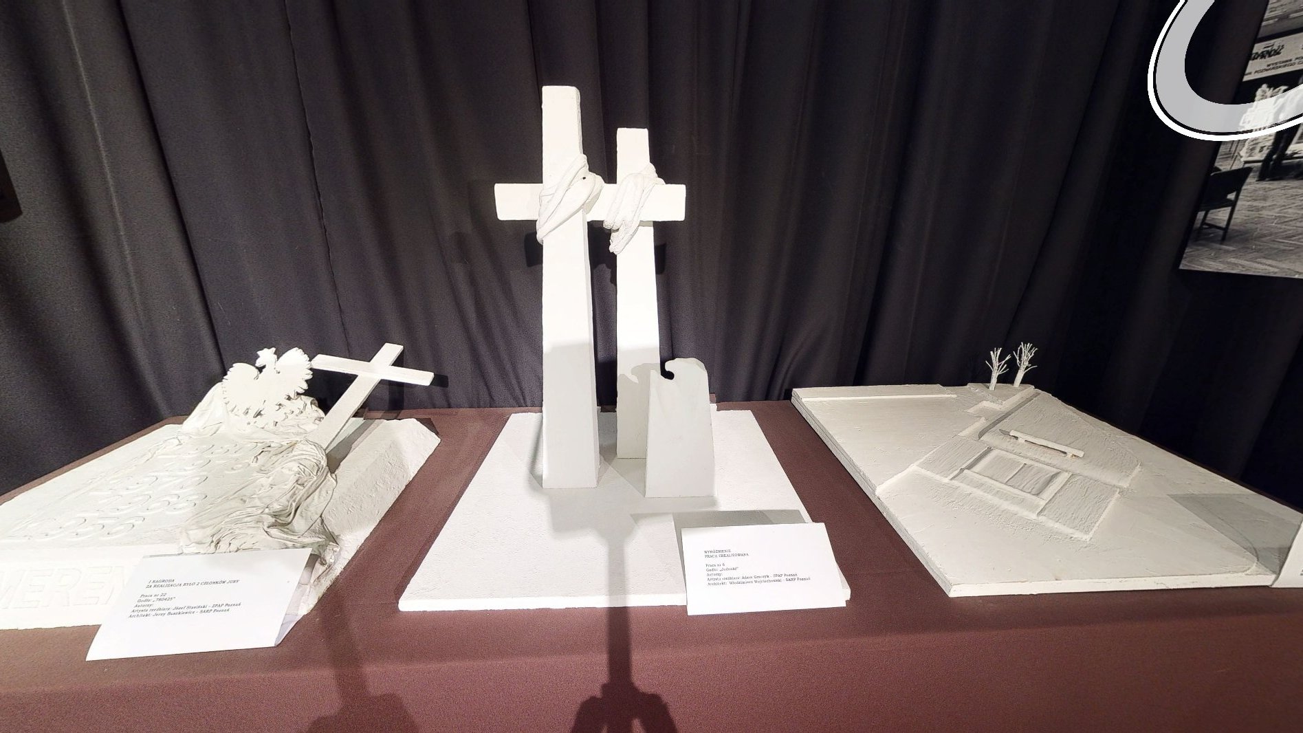 Biała makieta na stole. Przedstawia dwa krzyże obleczone sznurem i mniejszy pomnik, który przypomina orła. Przed makietą stoi tabliczka informacyjna.