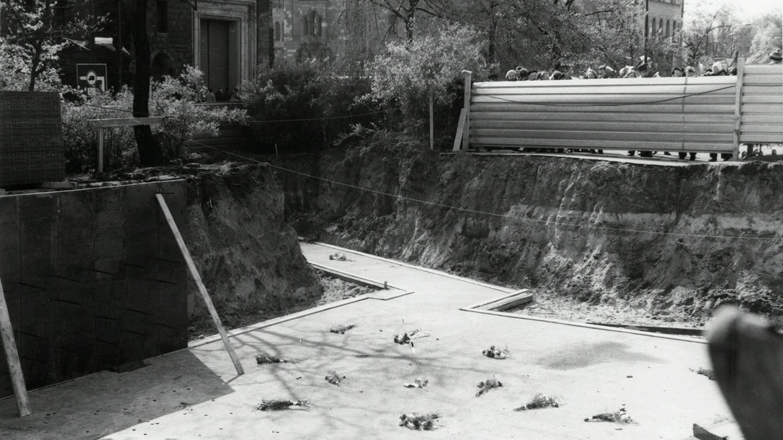 Czarno białe zdjęcie przedstawia wykopany dół. Na dnie znajdują się fundamenty pod pomnik. Zza płotu, który odgradza teren wychylają się mieszkańcy. W tle widać Filharmonię Poznańską.