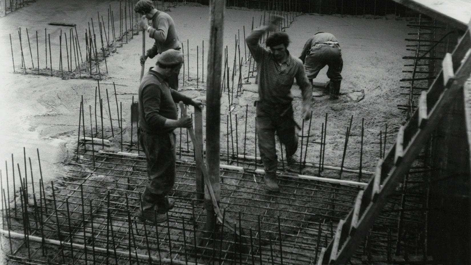 Czarno białe zdjęcie przedstawia pracowników, którzy przygotowują fundamenty pod pomnik. Czterech mężczyzn ubranych w stój roboczy montuje pionowe metalowe drążki.