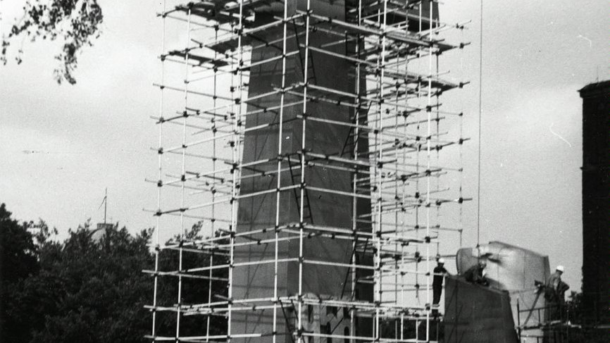 Czarno-białe zdjęcie przedstawia pomnik "Jedność". Wokół jednego z krzyży stoi rusztowanie. Pod rusztowaniem stoi dwóch mężczyzn. W prawym górnym rogu zwisa hak od dźwigu.