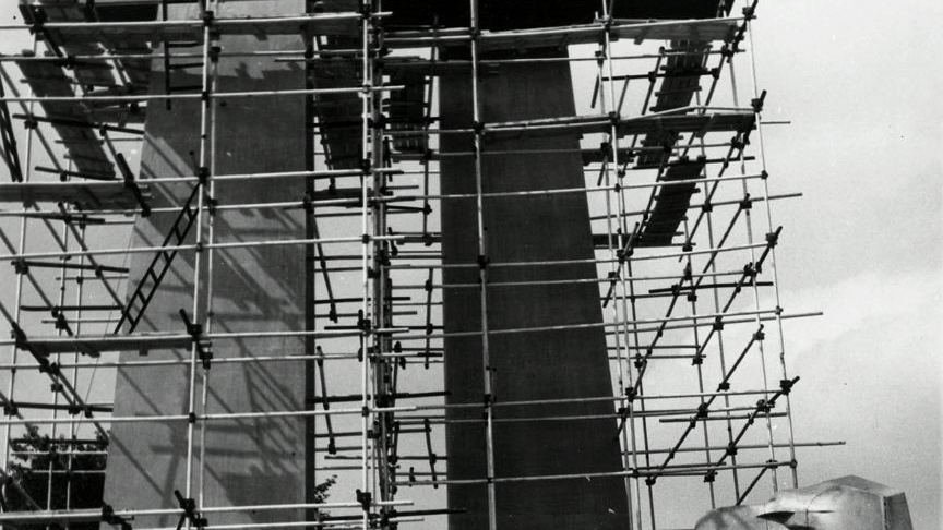 Czarno-białe zdjęcie przedstawia pomnik "Jedność". Pomniki dwóch krzyży i orła są otoczone rusztowaniami. Na jednym z nich stoi pracownik w stroju roboczym.