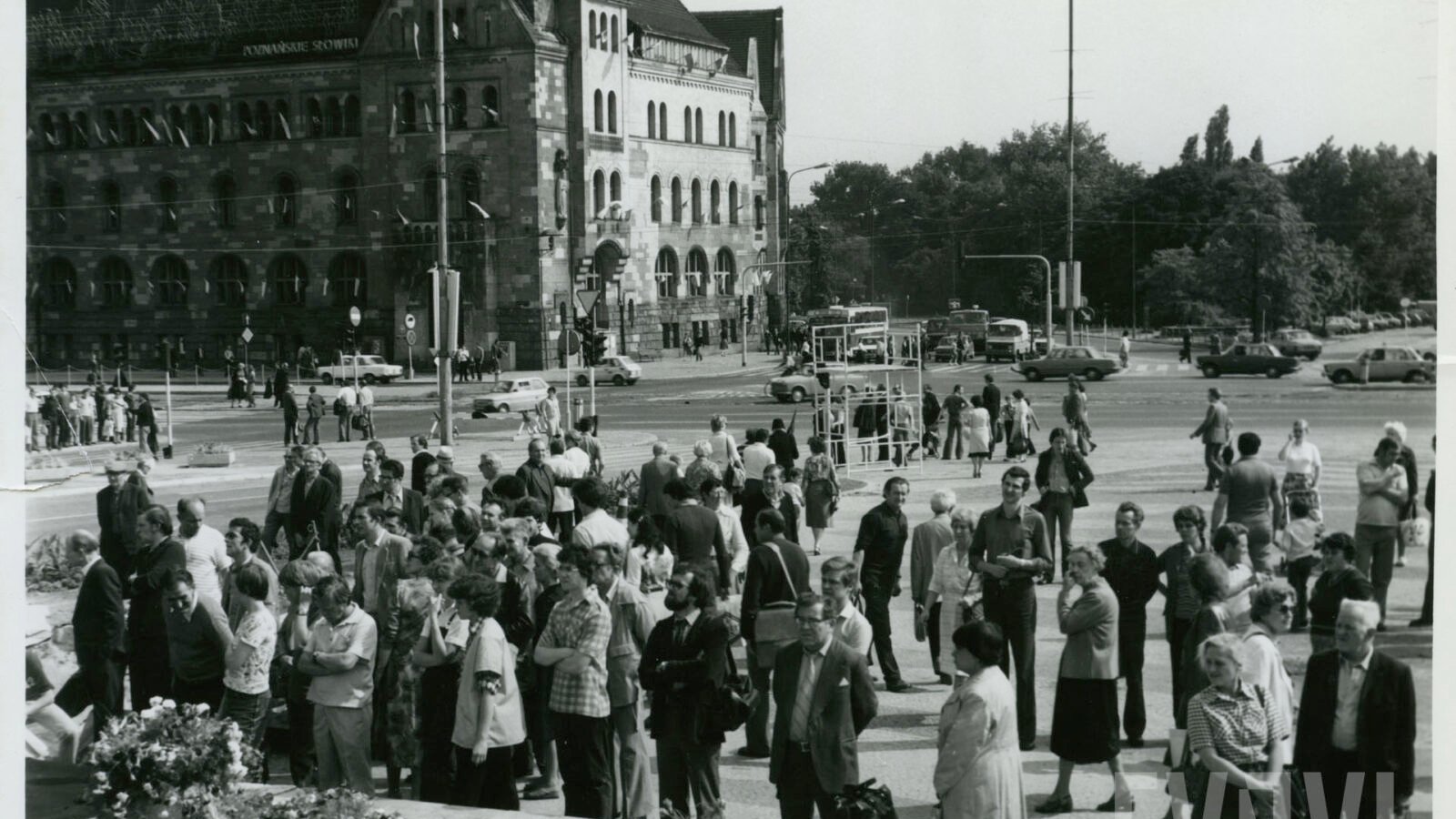 Czarno-białe zdjęcie przedstawia grupę mieszkańców, którzy oglądają budowę pomnia. Za nimi widać budynek Filharmonii Poznańskiej, park i ulicę.
