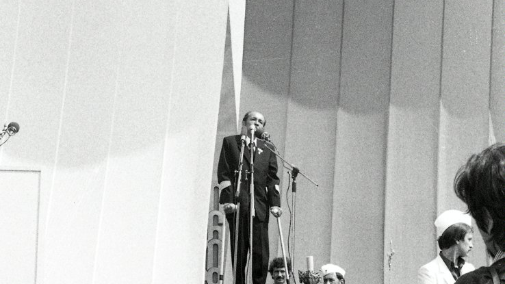 Czarno-biale zdjęcie. Na tle biało-czerwonych płacht na scenie przemawia Stanisław Matyja.