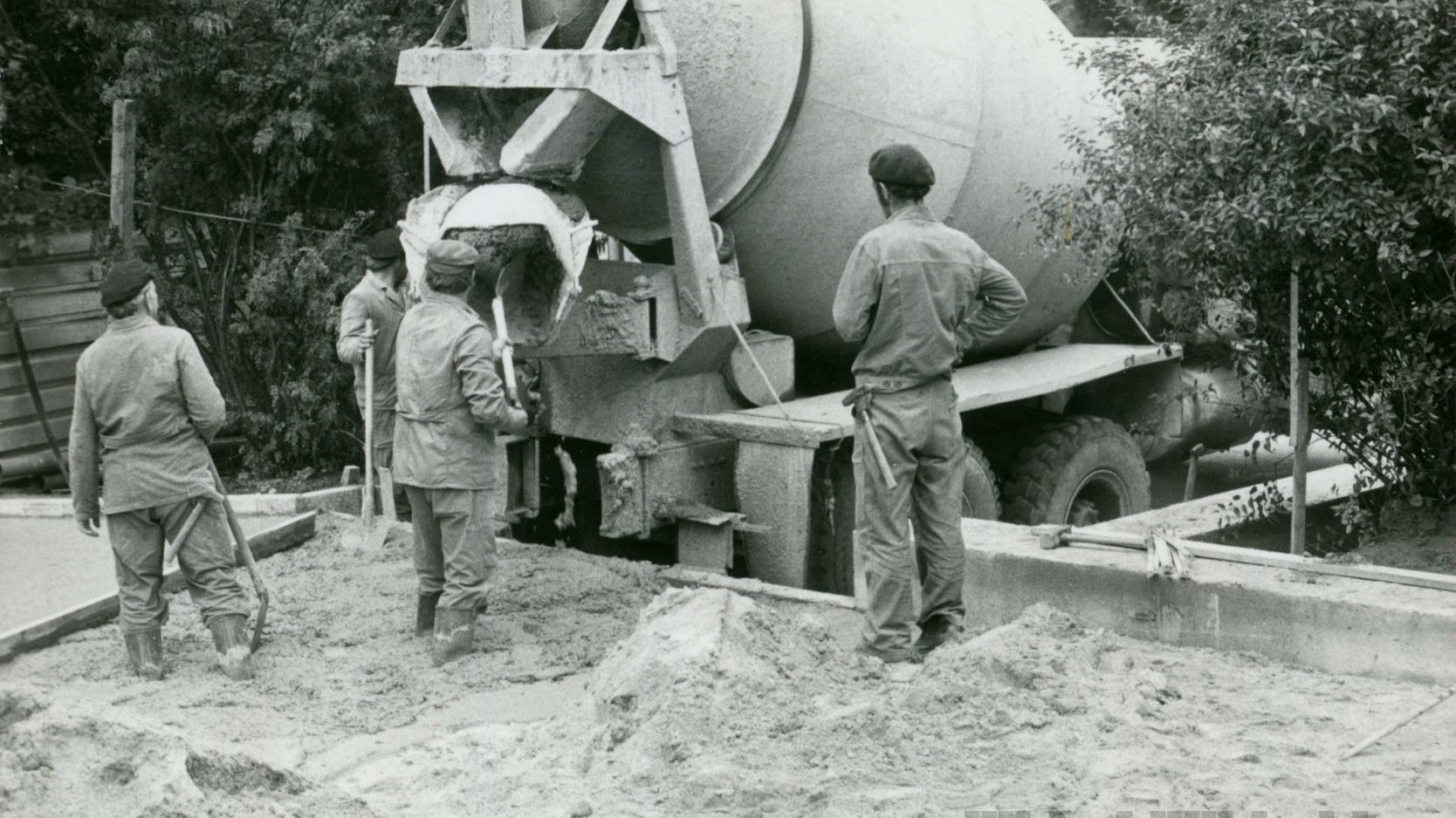 Czarno-białe zdjęcie. Czterech mężczyzn wylewa z betoniarki materiał do fundametu. Mężczyźni są ubrani w stroje robocze i mają w rękach łopaty.