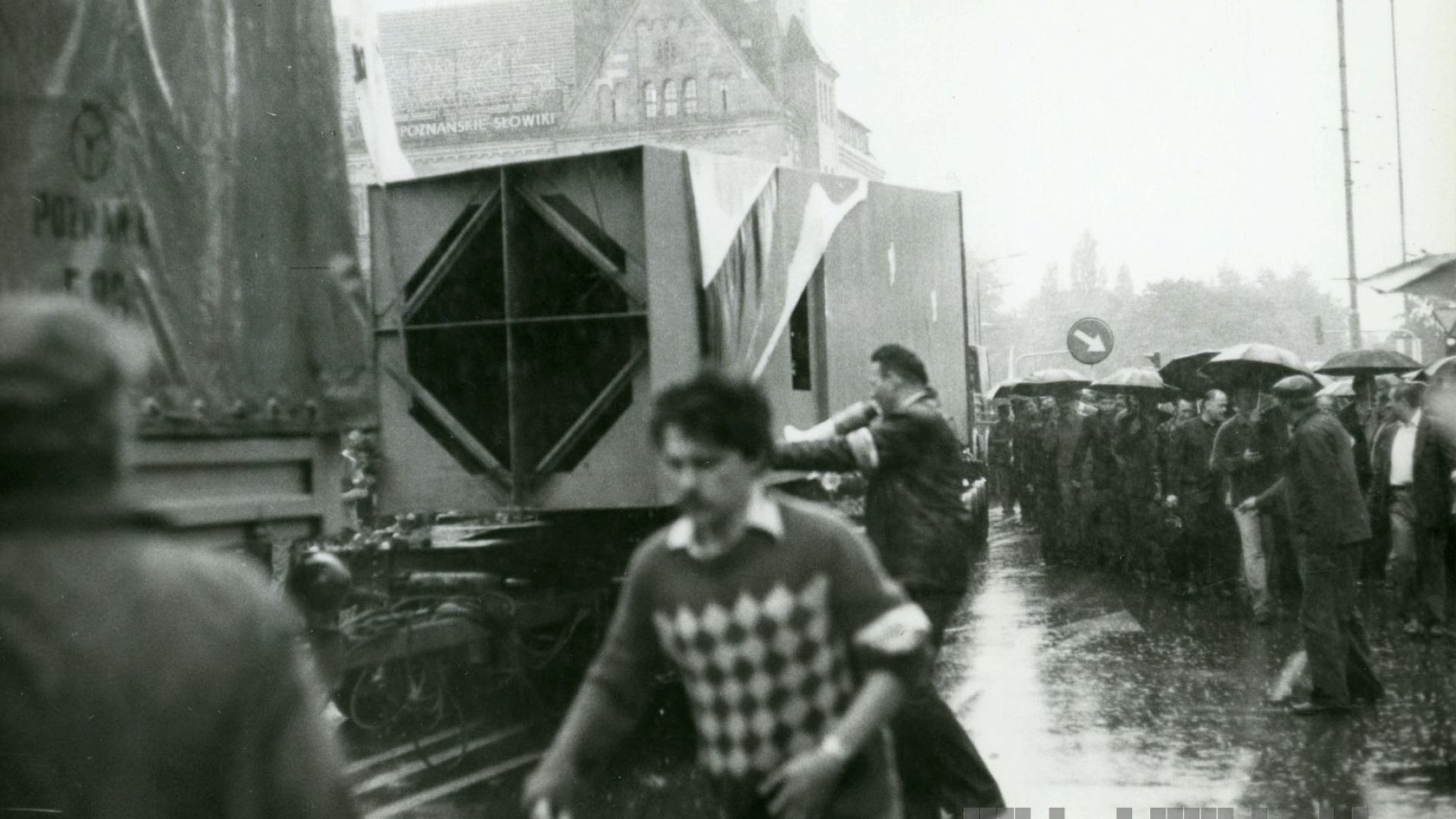 Czarno-białe zdjęcie przedstawia transport pomnika na ciężarówkach. Wokół samochodu zbiera się tłum, który trzyma parasolki. W oddali zamglony budynek Filharmonii Poznańskiej i park.