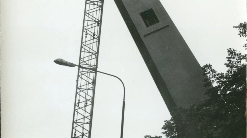 Czarno-białe zdjęcie przedstawia podnoszenie części pomnika przez dźwig. Pomnik jest pochylony nad drzewami w parku.