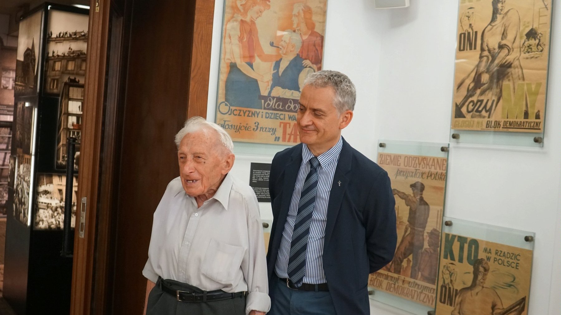 Dwóch mężczyzn. Po lewej starszy - Zenon Wechmann. Po prawej dyrektor muzeum. Mężczyźni stoją na tle plakatów propagandowych z okresu PRL-u.