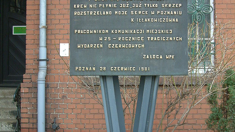 Metalowy pomnik, na którym znajduje się fragment wiersza Iłłakowiczówny. Pomnik składa się z dwóch szyn, które tworzą znak "V". Na nich przyczepiona jest prostokątna tablica z tekstem. W tle budynek i krzaki.