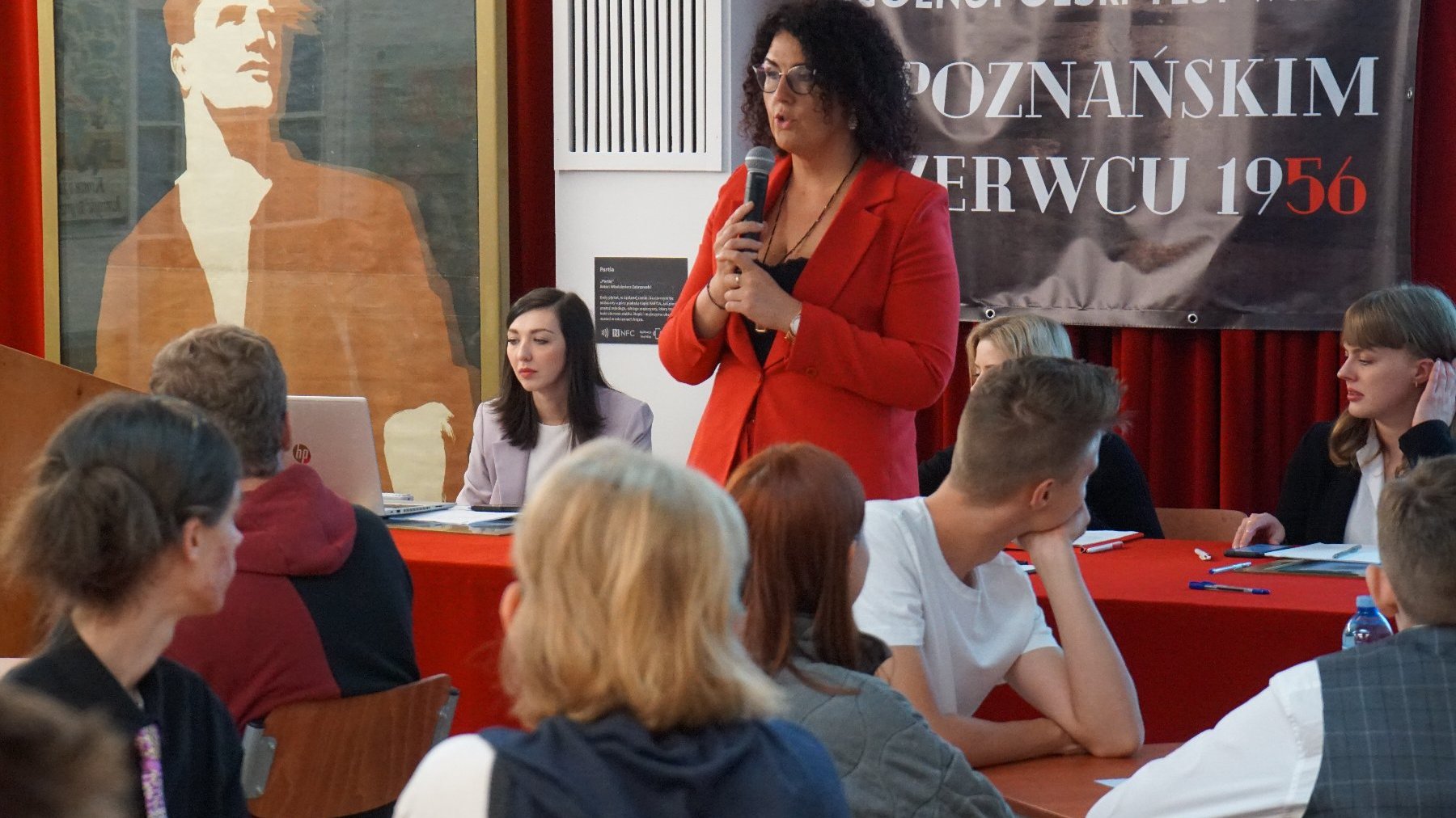 Kobieta z czarnymi włosami i w czerwonej marynarce przemawia przez mikrofon. Przed nią siedzą uczniowie. Za kobietą siedzą 3 kobiety. W tle plakat konkursowy.