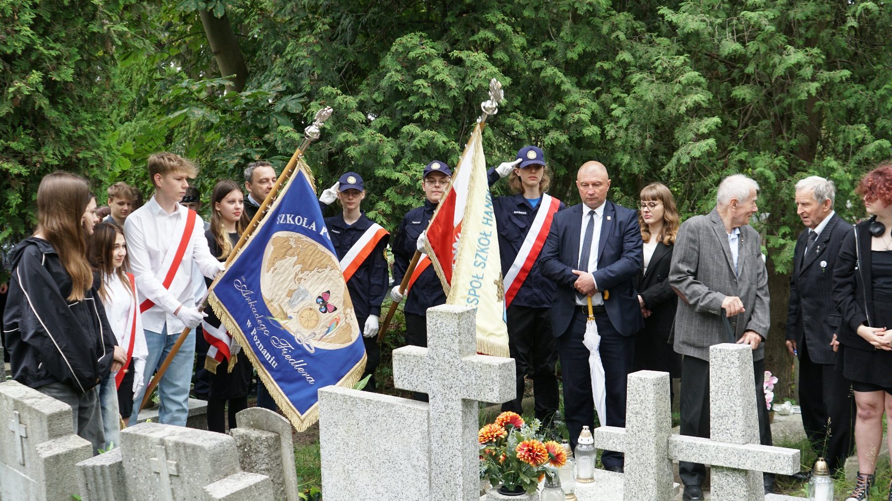 Zdjęcie przedstawia młodzież i dorosłych stojących przed grobami.