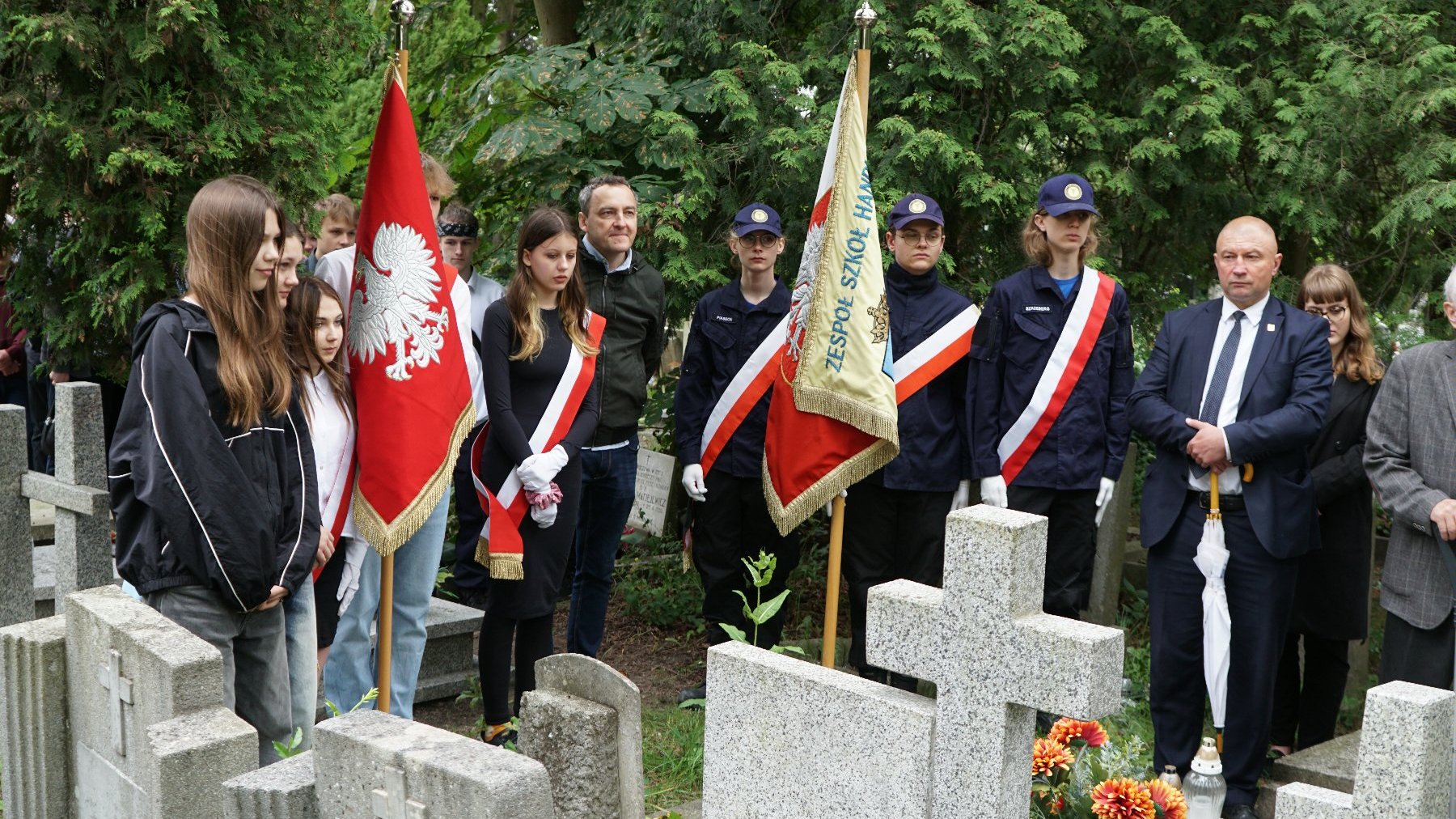 Zdjęcie przedstawia młodzież ze sztandarami stojącą przed grobami.